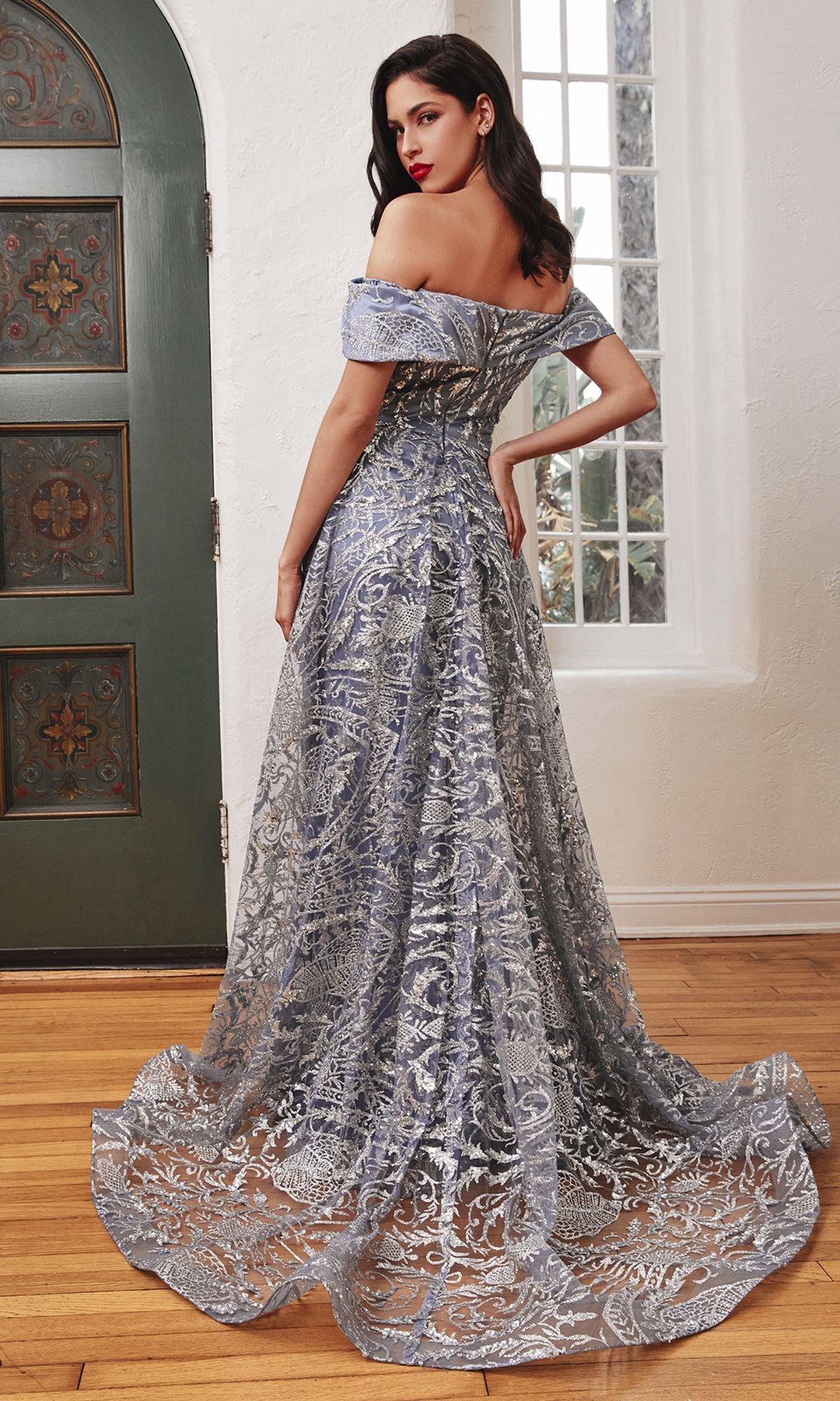 A woman wearing the Long Prom Dress with Glitter-Print Overskirt J836—an off-the-shoulder gown with silver and blue baroque glitter prints and a long train—stands indoors on a wooden floor near a window and green door, facing away.